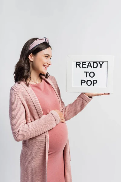 Joyful and pregnant woman looking at board with ready to pop lettering isolated on white — Stock Photo