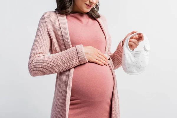 Vista recortada de la mujer embarazada sosteniendo pequeño babero bebé aislado en blanco - foto de stock