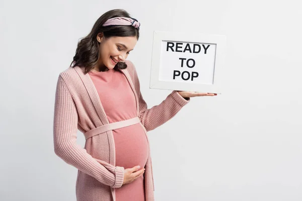 Mujer embarazada complacida sosteniendo el tablero con letras listas para estallar aisladas en blanco - foto de stock