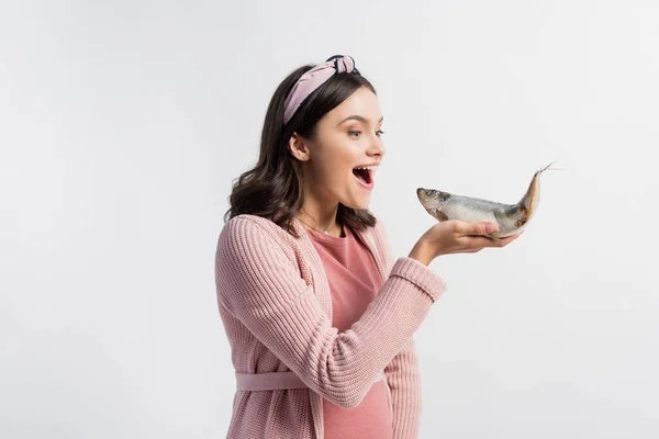 Mujer embarazada excitada con la boca abierta sosteniendo pescado seco aislado en blanco - foto de stock