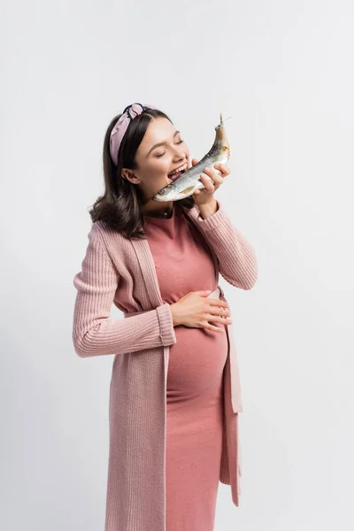 Беременная женщина с закрытыми глазами и открытым ртом ест сушеную рыбу, изолированную на белом — стоковое фото