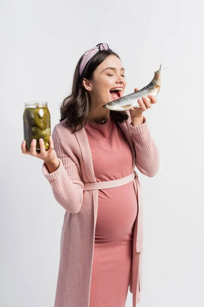 Mujer embarazada con boca abierta sosteniendo frasco con pepinos en escabeche y pescado seco aislado en blanco - foto de stock