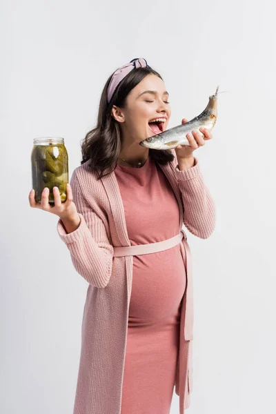 Pregnant woman with open mouth holding jar with pickled cucumbers and dried fish isolated on white — Stock Photo