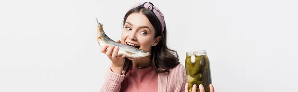 Panoramic shot of woman with open mouth holding jar with pickled cucumbers and dried fish isolated on white — Stock Photo