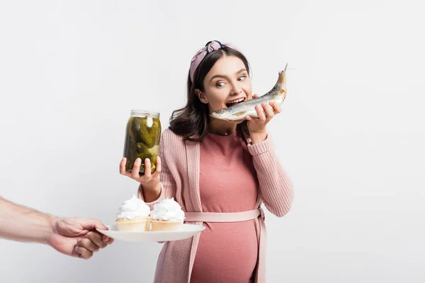 Mulher grávida segurando frasco com pepinos em conserva e comendo peixe seco enquanto olha para cupcakes na placa na mão masculina isolado no branco — Fotografia de Stock