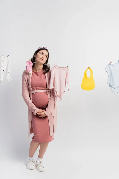 Upset and pregnant woman touching belly near baby clothes on clothing line on white — Stock Photo