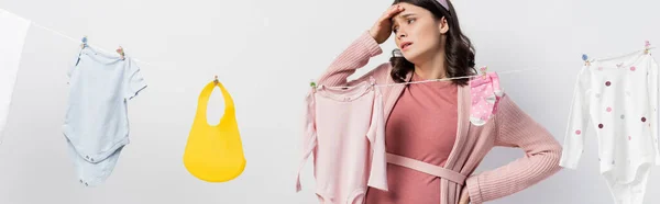 Imagen horizontal de la mujer embarazada triste de pie con la mano en la cadera cerca de la ropa de bebé en blanco - foto de stock