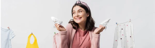 Plan panoramique de la femme enceinte tenant des bottes minuscules près des vêtements de bébé sur la ligne de vêtements isolés sur blanc — Photo de stock