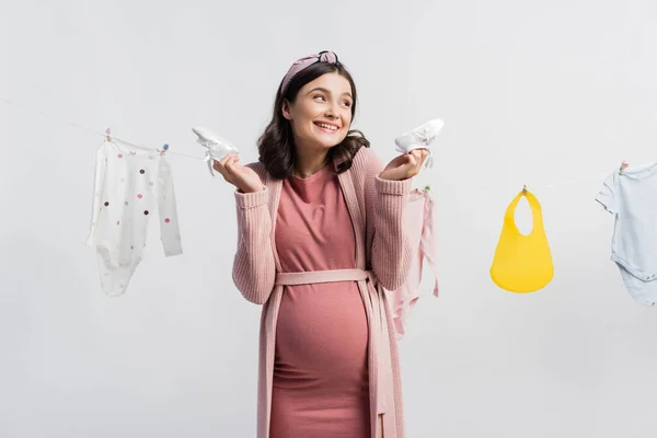 Gioiosa donna incinta che tiene piccoli stivali vicino a vestiti per bambini sulla linea di abbigliamento isolata su bianco — Foto stock