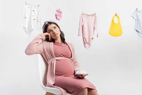 Femme enceinte coûteuse tenant des bottes minuscules et assis près de vêtements de bébé sur la ligne de vêtements isolés sur blanc — Photo de stock