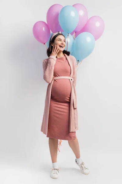 Donna gioiosa e incinta guardando lontano mentre in piedi vicino palloncini su bianco — Foto stock