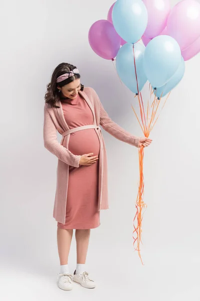 Mulher alegre e grávida no vestido tocando barriga e segurando balões em branco — Fotografia de Stock