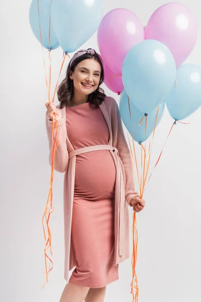 Joyful and pregnant woman in pink dress holding balloons on white — Stock Photo