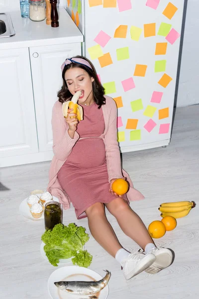 Pregnant woman eating banana near jar with canned cucumbers, cupcakes and oranges while sitting on floor — Stock Photo