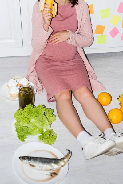 Partial view of pregnant woman holding banana near jar with canned cucumbers, cupcakes and oranges while sitting on floor — Stock Photo