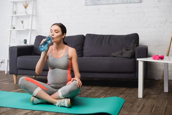 Mujer embarazada sentada en la alfombra de fitness mientras bebe agua de la botella de deportes - foto de stock