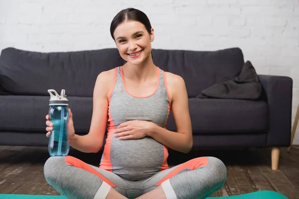 Joyful pregnant woman sitting with sports bottle at home — Stock Photo