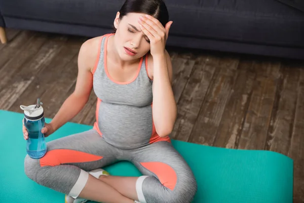Высокий угол зрения уставшей беременной женщины, держащей спортивную бутылку и сидящей на коврике — стоковое фото
