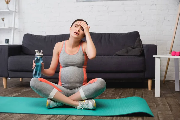 Tired pregnant woman holding sports bottle and sitting on fitness mat — Stock Photo