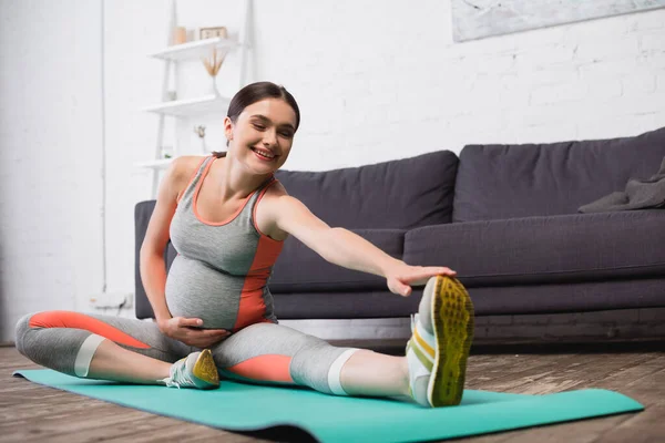 Mulher grávida satisfeito em sportswear alongamento no tapete de fitness — Fotografia de Stock