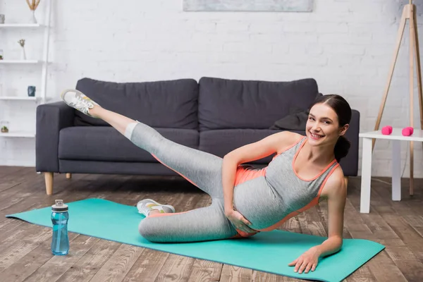 Mulher grávida satisfeito em sportswear alongamento no tapete de fitness perto de garrafa de esportes — Fotografia de Stock