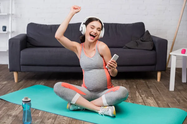 Mujer embarazada emocionada en auriculares inalámbricos escuchando música y sosteniendo el teléfono inteligente - foto de stock