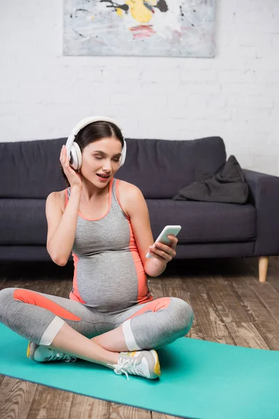 Беременная женщина в беспроводных наушниках слушает музыку и держит смартфон — стоковое фото