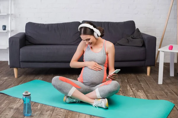 Joyeuse femme enceinte dans les écouteurs sans fil écouter de la musique et tenant smartphone tout en étant assis sur tapis de fitness — Photo de stock