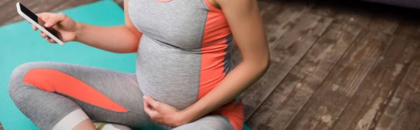 Панорамный урожай беременной женщины, держащей смартфон, сидящей на фитнес-коврике — стоковое фото
