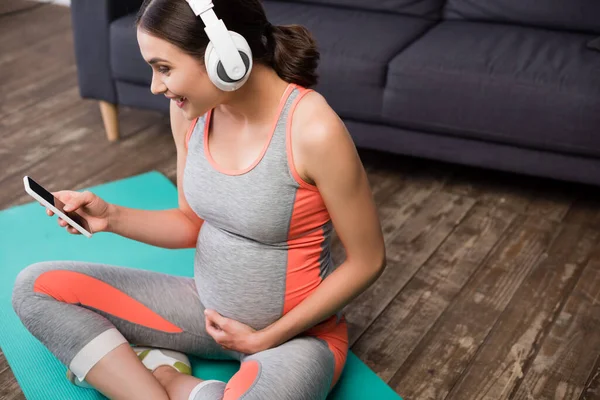 Радостная беременная женщина в беспроводных наушниках слушает музыку и использует смартфон, сидя на фитнес-коврик — стоковое фото