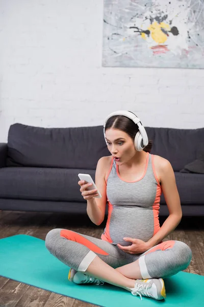 Mujer sorprendida y embarazada en auriculares inalámbricos escuchando música y utilizando el teléfono inteligente mientras se sienta en la alfombra de fitness - foto de stock