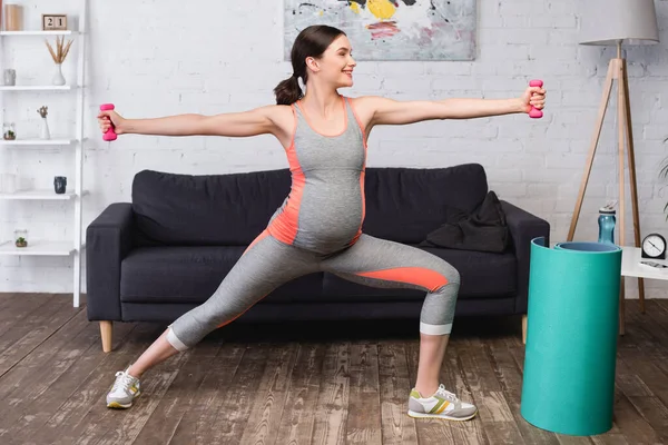 Gioiosa donna incinta in piedi con le mani tese durante l'esercizio con manubri rosa a casa — Foto stock