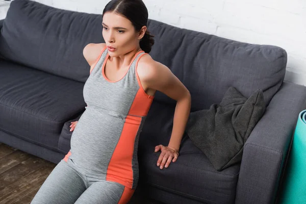 Morena e mulher grávida no sportswear respiração durante o exercício perto do sofá na sala de estar — Fotografia de Stock