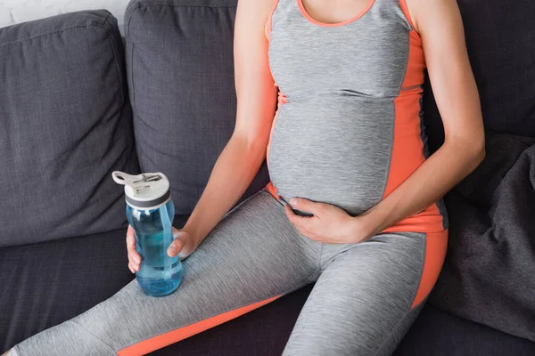 Cropped view of pregnant woman sitting on sofa and holding sports bottle with water — Stock Photo