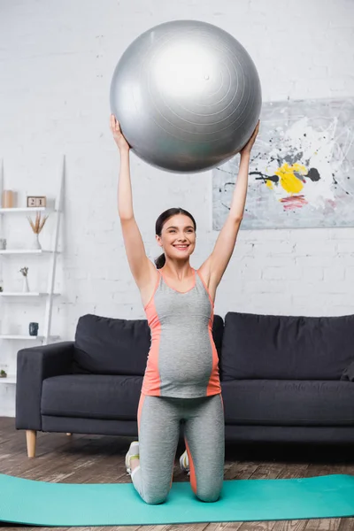 Mujer alegre y embarazada haciendo ejercicio con la pelota de fitness en la sala de estar - foto de stock