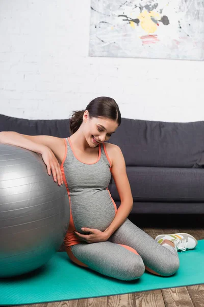 Pleased and pregnant woman touching belly while sitting near fitness ball in living room — Stock Photo