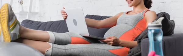 Спортивна та вагітна жінка використовує ноутбук біля спортивної пляшки та гантелі — стокове фото