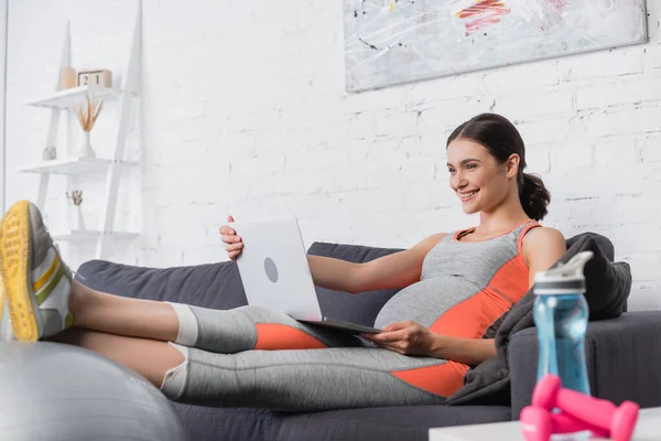 Sportive and pregnant woman looking at laptop near sports bottle and dumbbells — Stock Photo