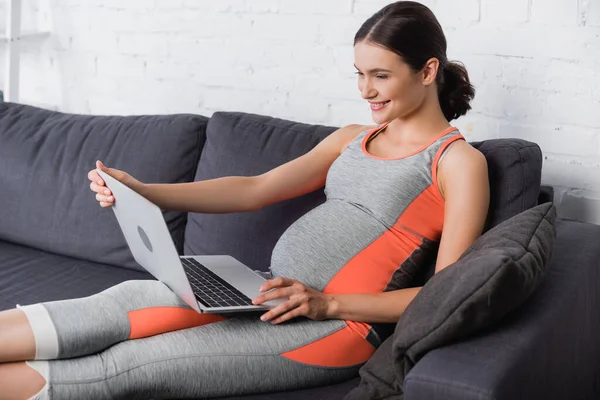 Mujer alegre, deportiva y embarazada usando el ordenador portátil en casa - foto de stock