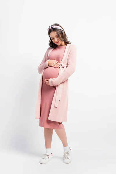 Full length view of pregnant woman in cardigan and headband touching belly on white — Stock Photo