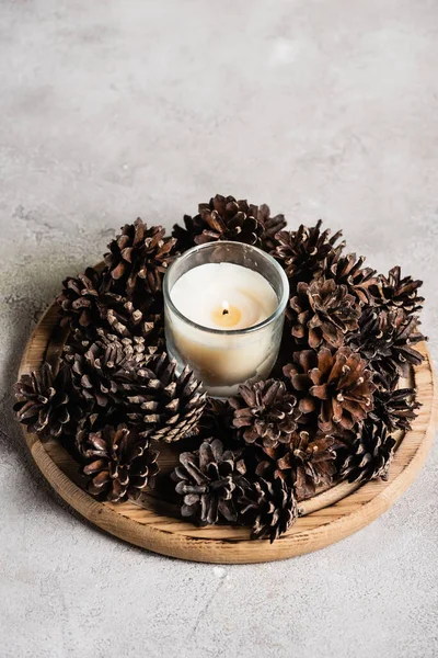 Scented candle with pine cones on wooden plate on grey and textured background — Stock Photo