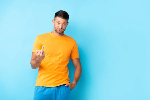 Homme en t-shirt debout avec la main dans la poche et montrant majeur sur bleu — Photo de stock