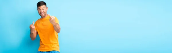 Smiling man showing middle fingers and looking at camera on blue, banner — Stock Photo