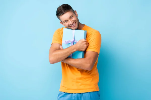 Homme heureux en t-shirt embrassant boîte cadeau enveloppé sur bleu — Photo de stock