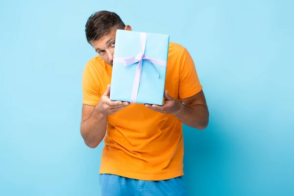 Чоловік у футболці ховається за загорнутою подарунковою коробкою на синьому — стокове фото