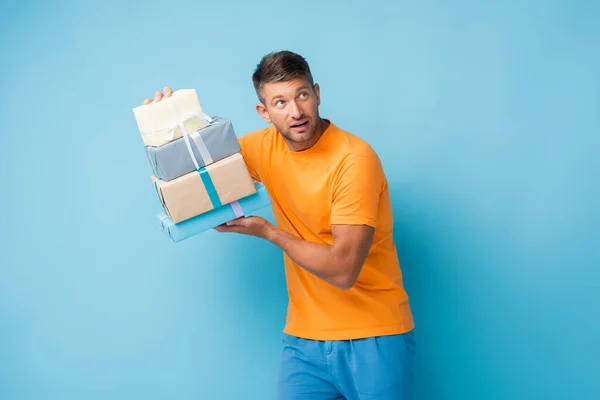 Curioso hombre en camiseta sosteniendo regalos envueltos y mirando hacia otro lado en azul — Stock Photo