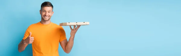 Веселий чоловік у футболці тримає картонну коробку для піци і показує великий палець на синьому, банер — стокове фото
