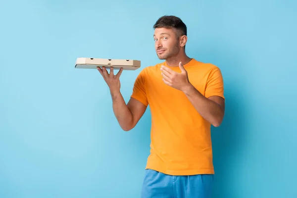 Hombre en camiseta sosteniendo caja de pizza de cartón y mirando a la cámara en azul - foto de stock