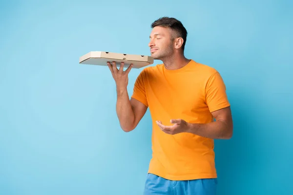 Homme en t-shirt tenant et sentant boîte à pizza carton sur bleu — Photo de stock