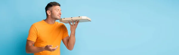 Hombre en camiseta sosteniendo y oliendo caja de pizza de cartón en azul, bandera - foto de stock